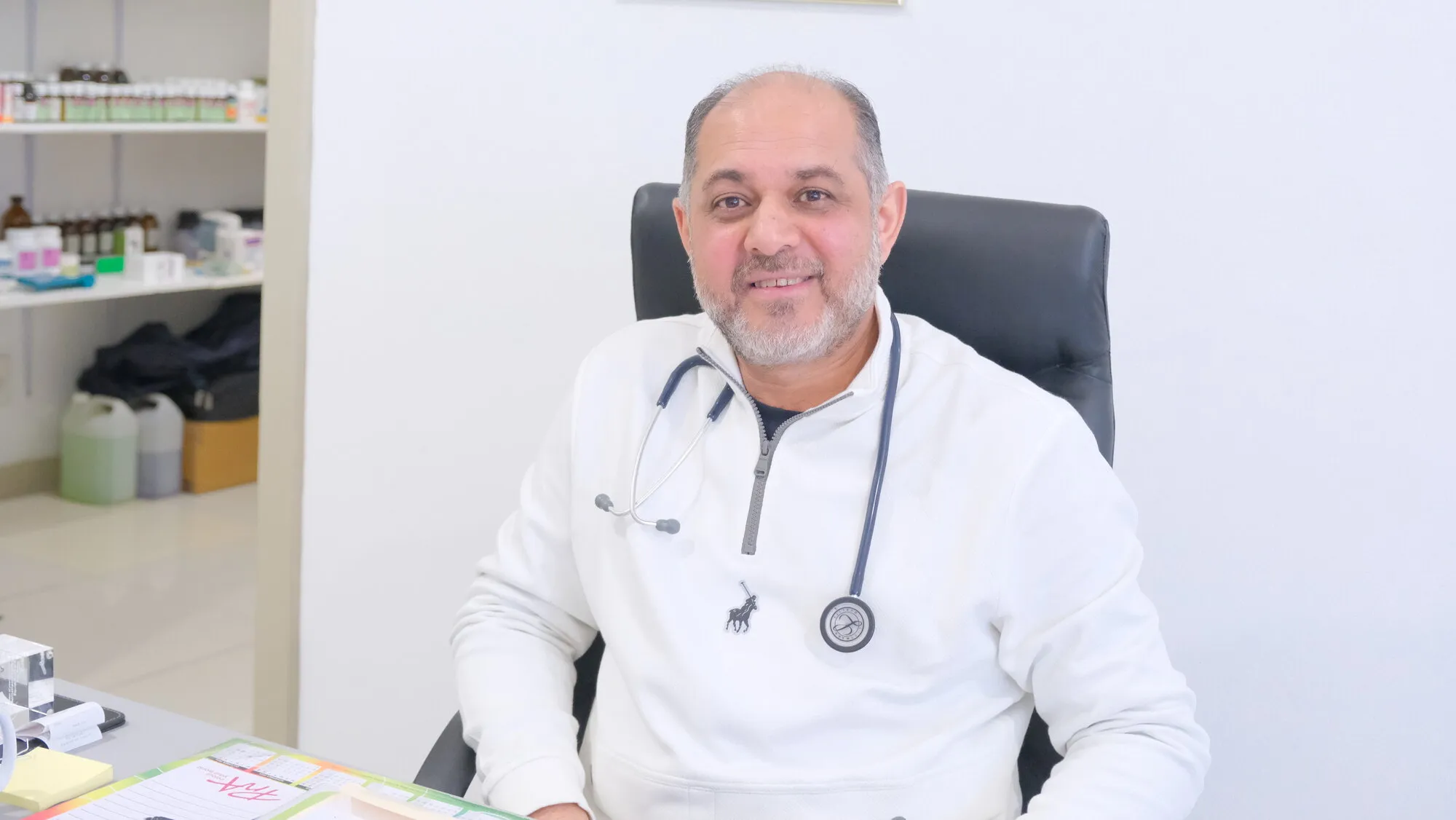 Dr MS Nalla seated at his desk in the consulting room, wearing a stethoscope, with dispensary shelves behind him