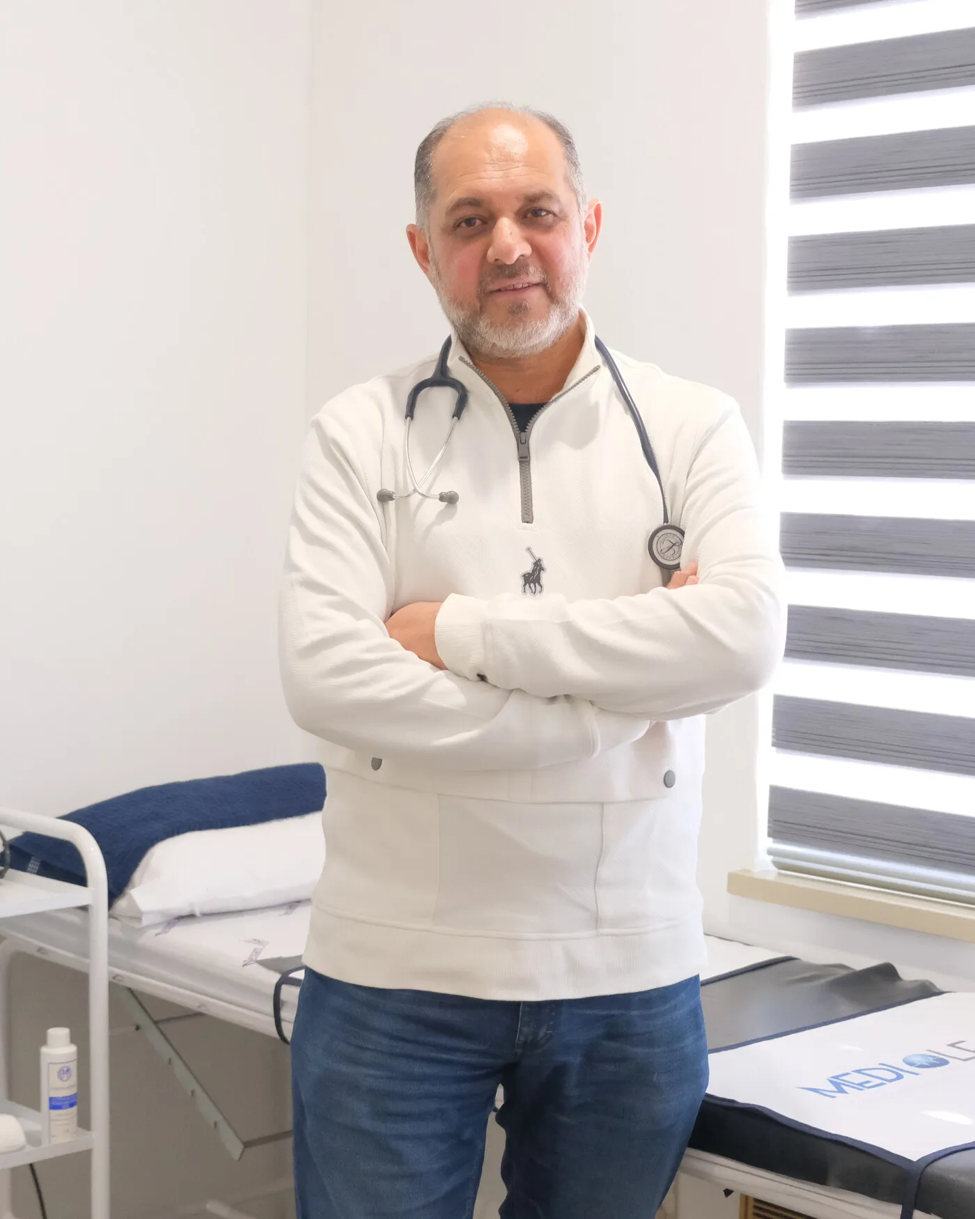 Dr MS Nalla standing in the treatment room with his arms folded, wearing a stethoscope, beside the examination couch