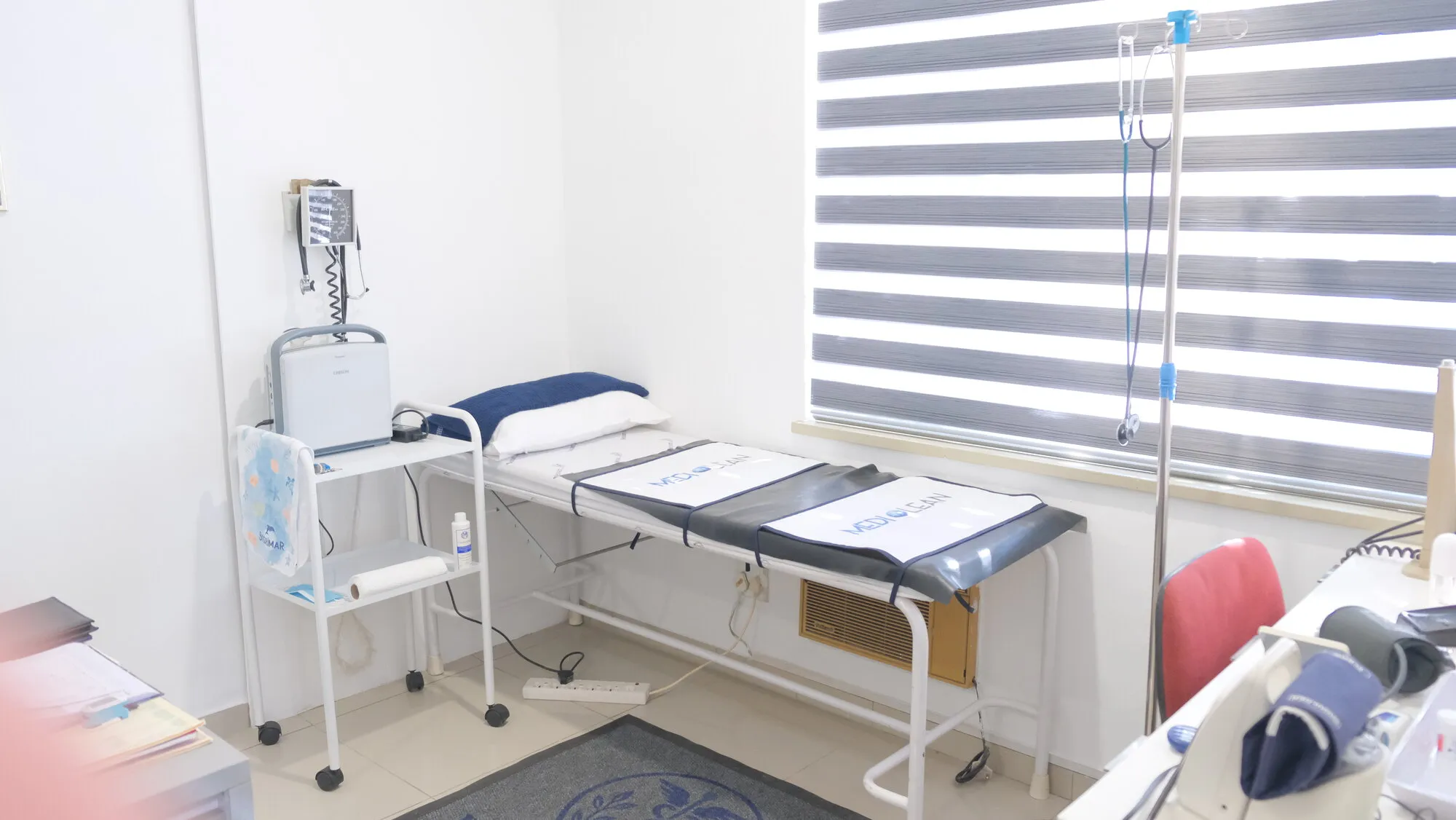 The treatment room, with an examination couch under the window, a wall mounted blood pressure monitor and a drip stand